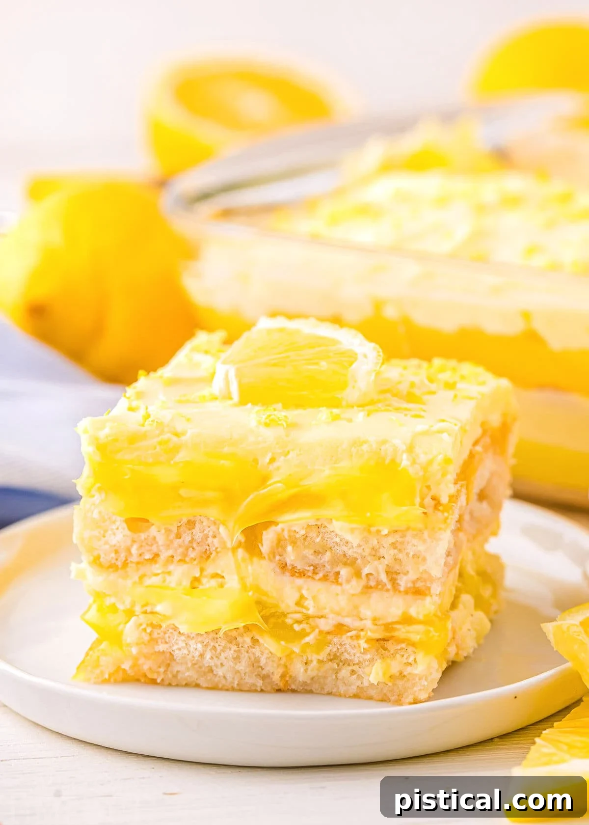 A perfectly portioned square of layered lemon tiramisu dessert on a white plate, with the rest of the dessert visible in the background, showcasing its vibrant layers.