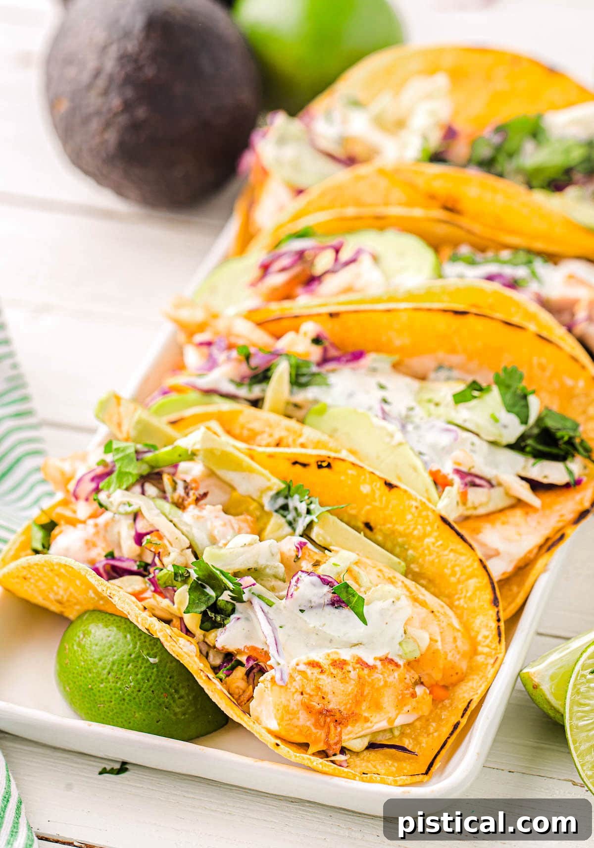 Easy Crispy Fish Tacos 2 Three perfectly assembled fish tacos on a white platter, drizzled with creamy fish taco sauce and garnished with fresh cilantro. Lime wedges are placed alongside, with a hint of avocado in the background, ready to elevate this quick and easy weeknight dinner.