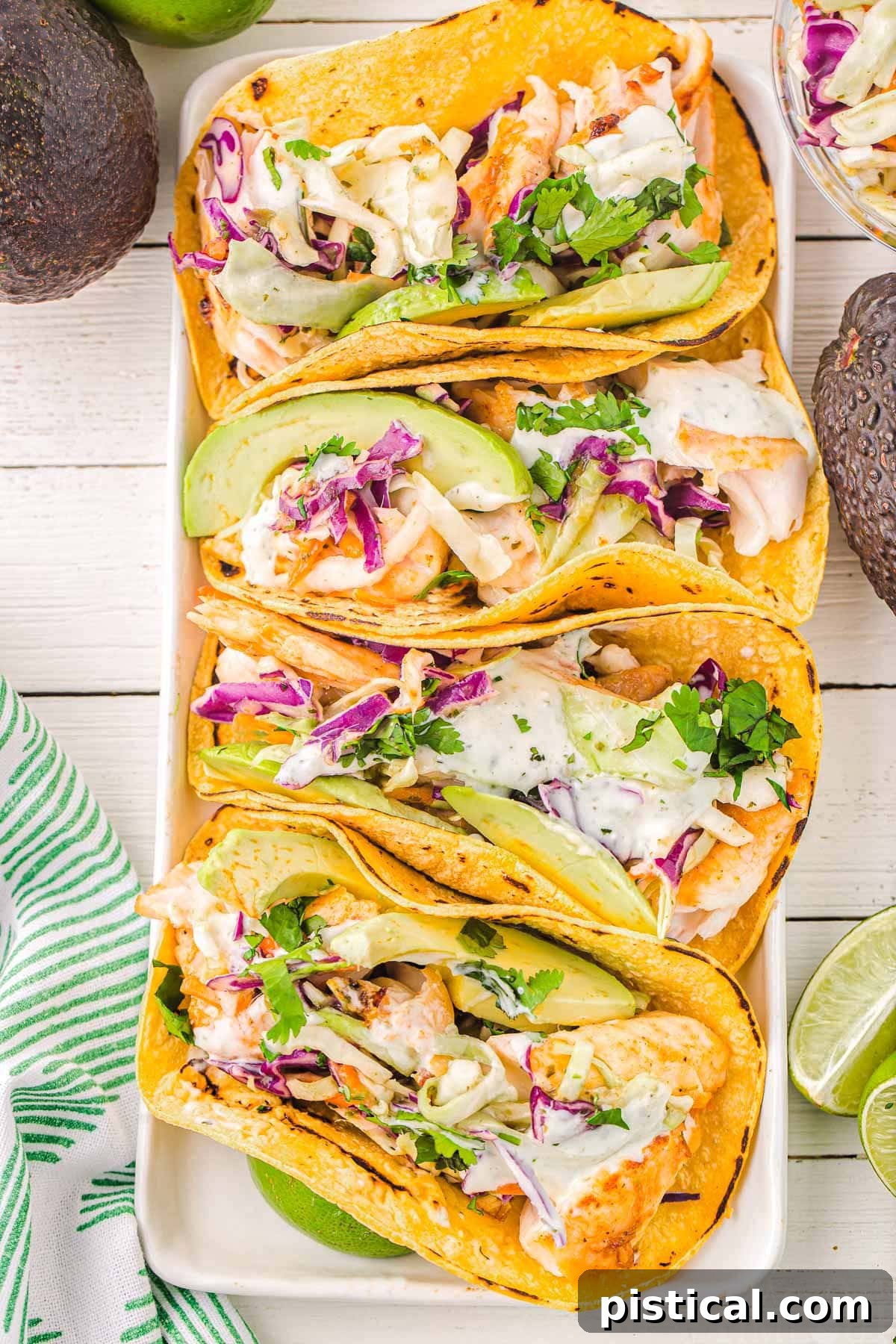 Easy Crispy Fish Tacos 3 A top-down view of four perfectly arranged fish tacos on a white rectangular plate, garnished with vibrant avocados, fresh cilantro, and bright lime wedges, showcasing a complete and appealing meal.