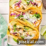 Top down view of four fish tacos on a white rectangle plate with avocados and cilantro and lime wedges around the plate.