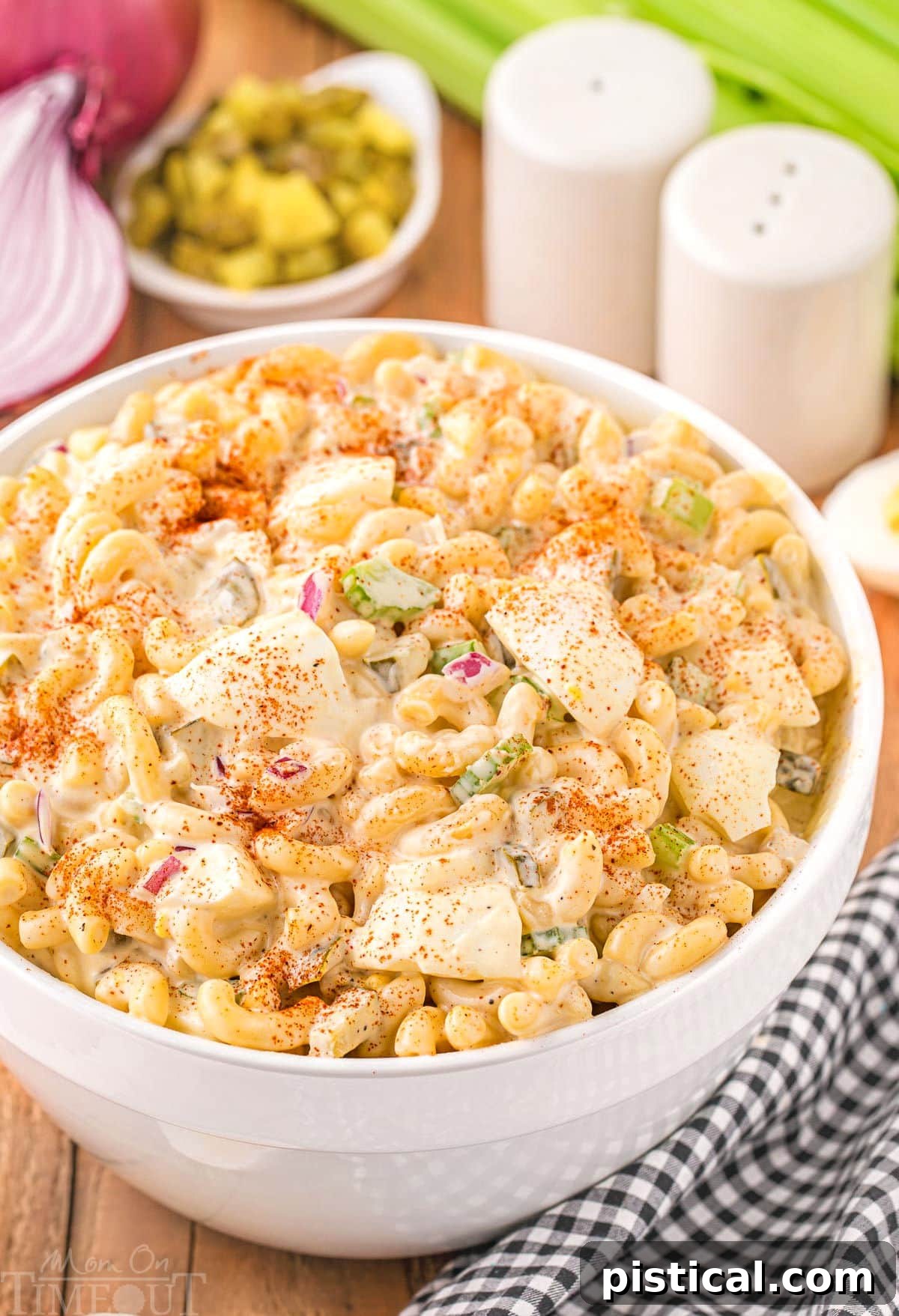A generous serving of deviled egg macaroni salad in a white bowl, garnished with a sprinkle of paprika. Sliced red onion, pickles, and celery are visible in the background, hinting at the fresh ingredients within.