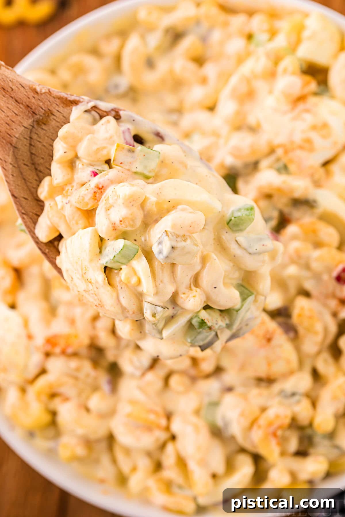 A wooden spoon lifting a generous scoop of creamy deviled egg macaroni salad from a large white serving bowl, showcasing the rich texture and colorful mix-ins.