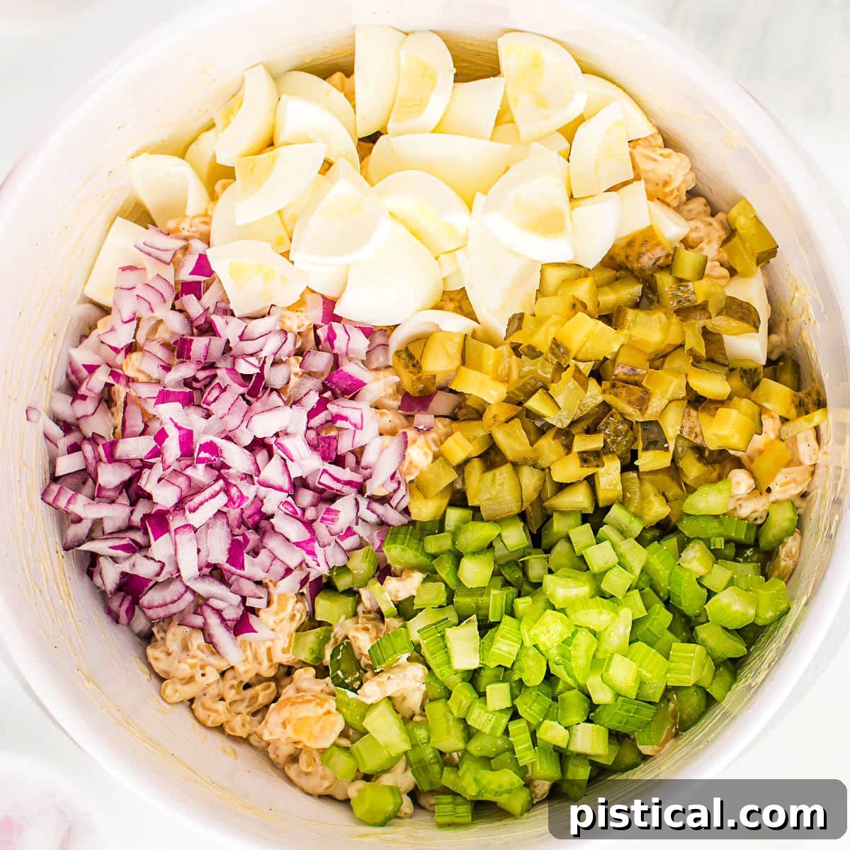A top-down view of a white bowl filled with cooked macaroni, and separate piles of chopped hard-boiled egg whites, pickles, celery, and red onion, ready to be combined with the dressing.