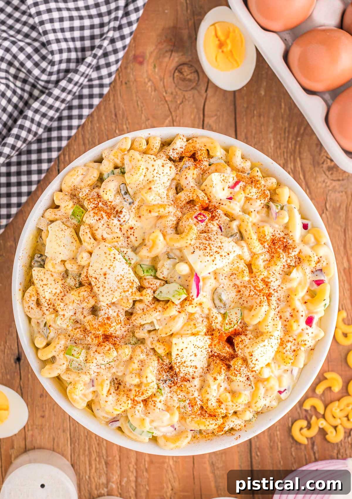 A large white serving bowl filled with deviled egg macaroni salad, set on a rustic wooden backdrop. Whole and sliced hard-boiled eggs are scattered around the bowl, alongside a white and black checkered towel, creating an inviting rustic scene.