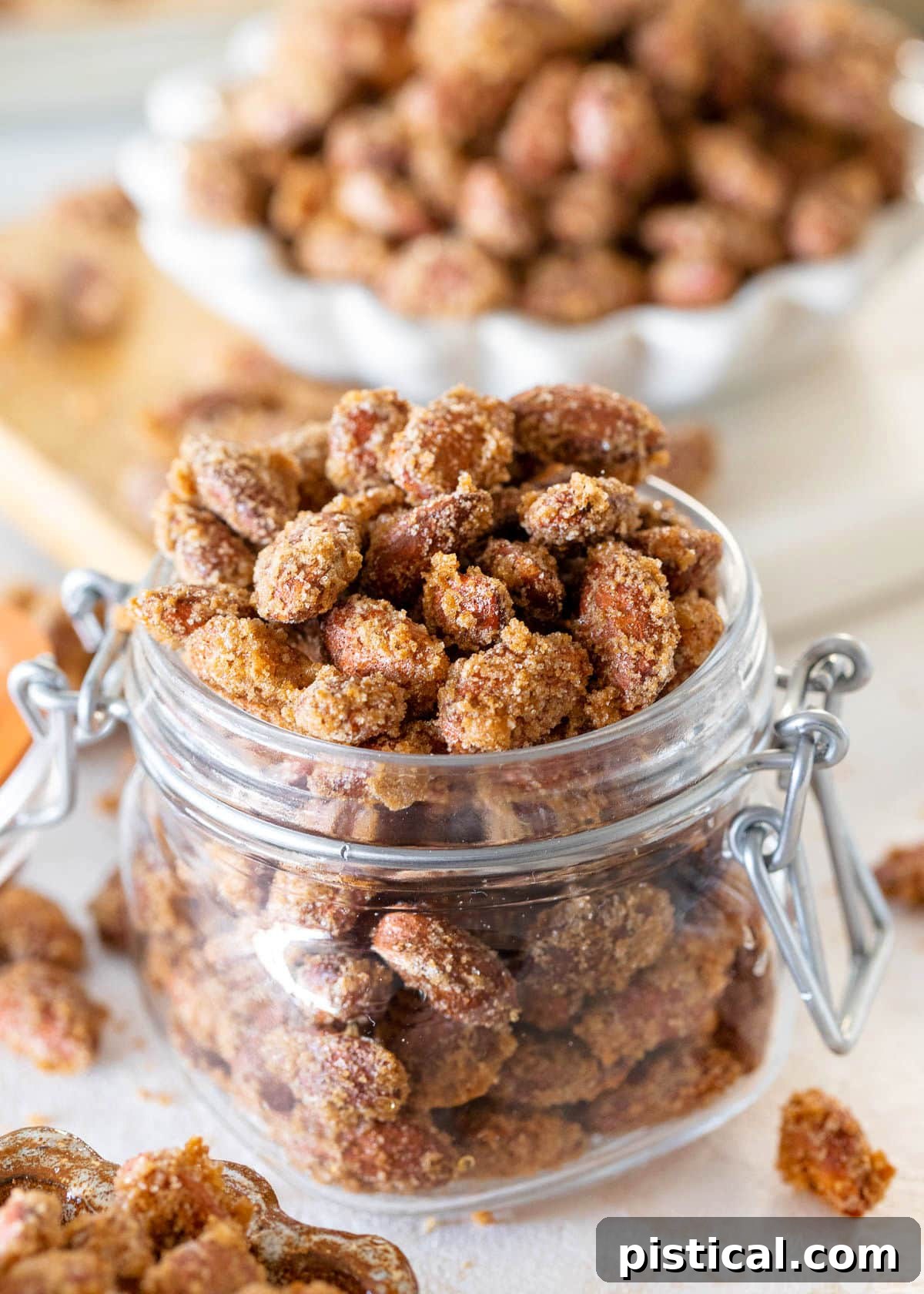 A glass jar overflowing with candied almonds, with a small white bowl of candied almonds in the background.
