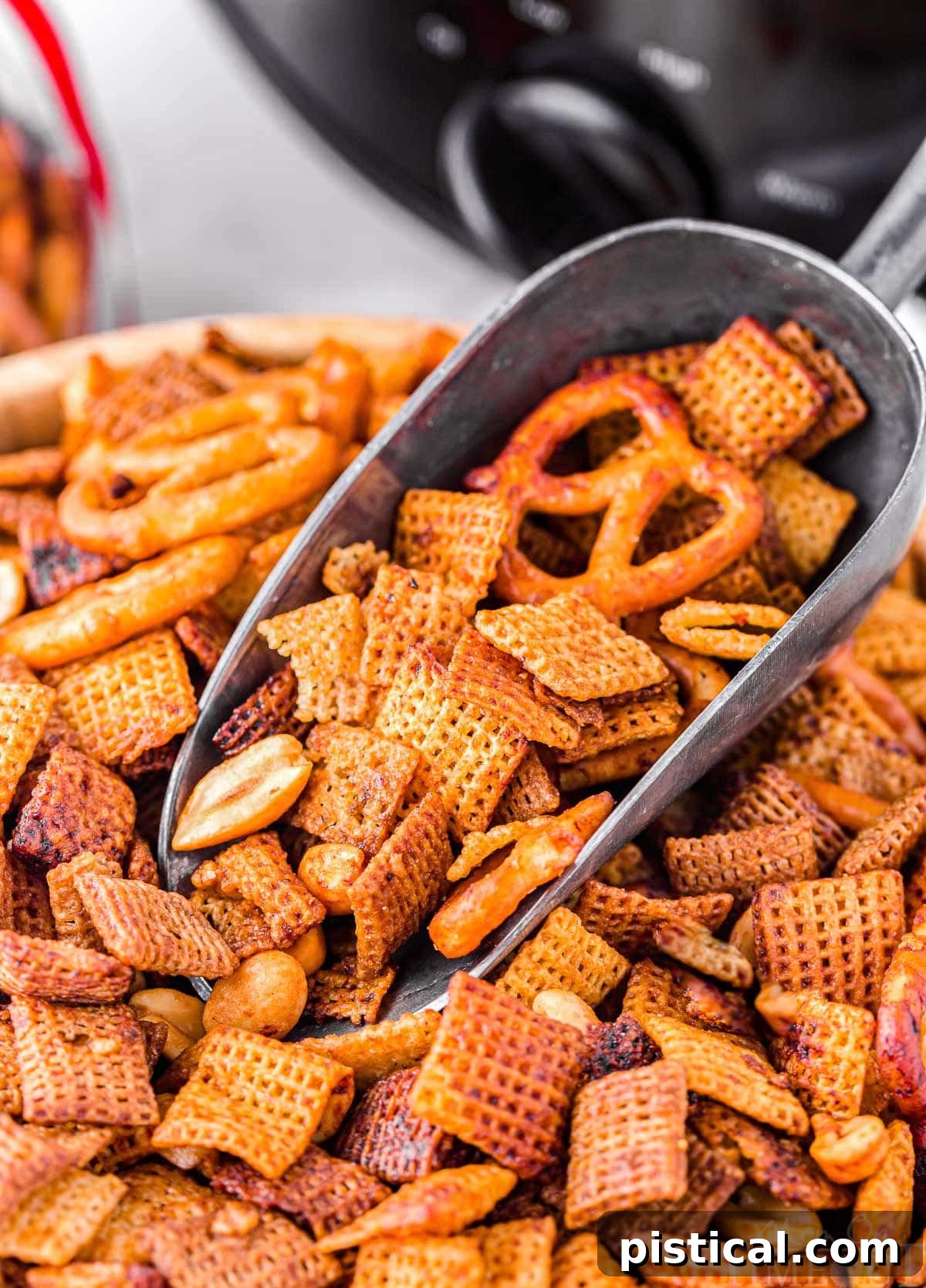 Large wooden bowl brimming with golden-brown slow cooker Chex Mix, with a metal scoop dipping in. A black slow cooker is visible in the background.