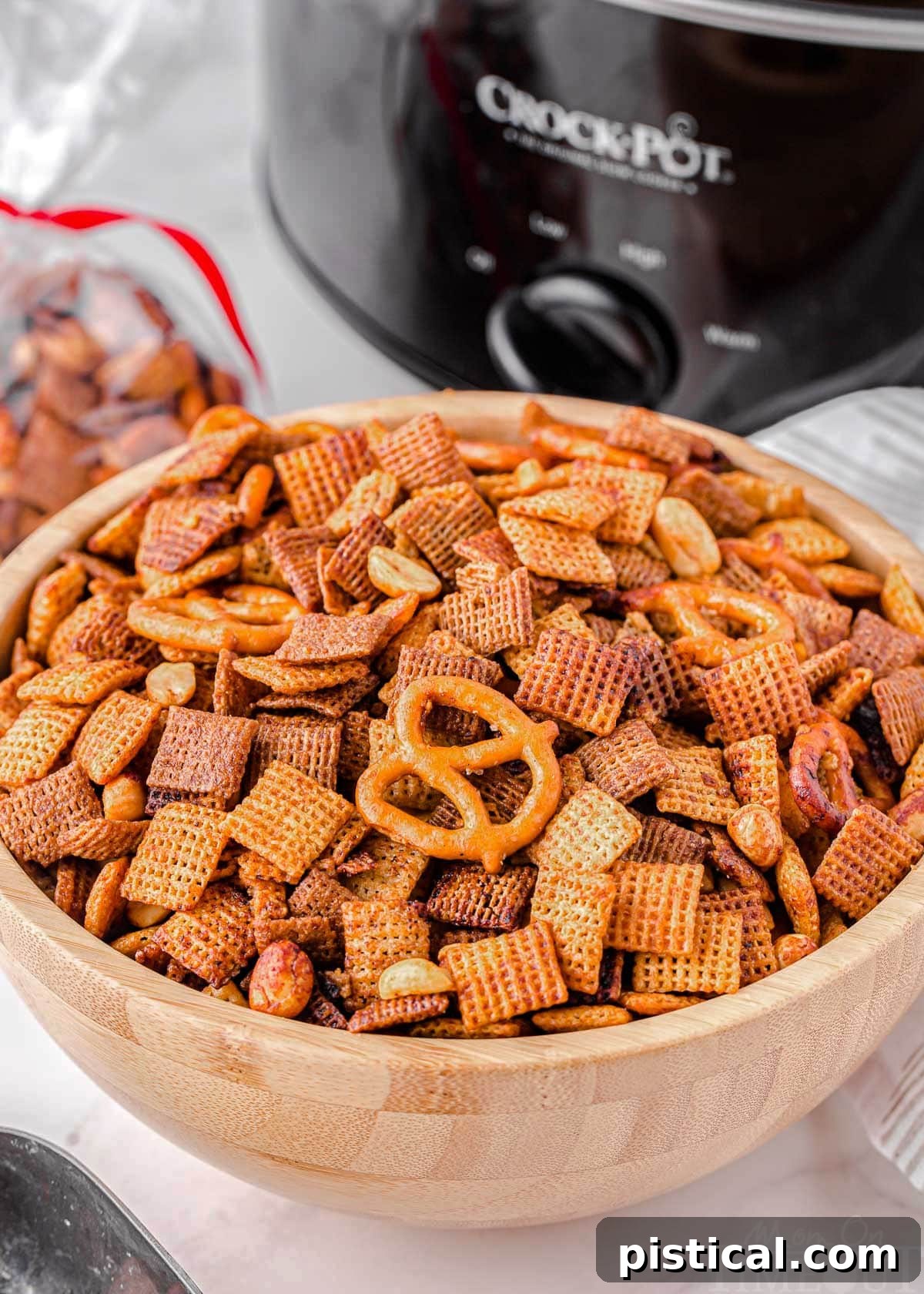 A wooden bowl filled with Crockpot Chex Mix positioned in front of a black slow cooker, showcasing the homemade snack.