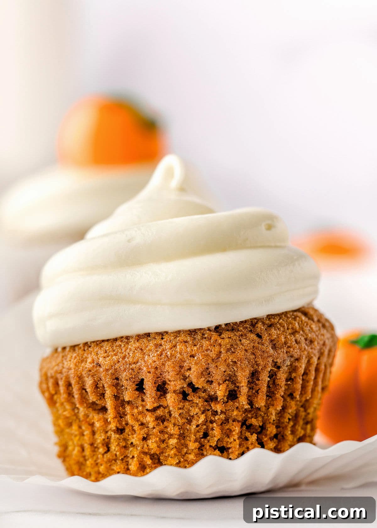 Decadent Pumpkin Cupcakes with Silky Cream Cheese Frosting 3 A beautifully frosted pumpkin cupcake, with its liner partly peeled back, sitting among other blurred cupcakes in the background.