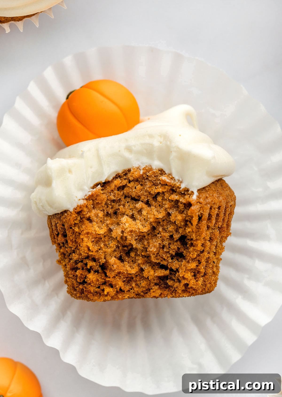 Decadent Pumpkin Cupcakes with Silky Cream Cheese Frosting 7 A top-down shot of a pumpkin cupcake lying on its side, showing its moist and tender crumb from a bite taken out. It's resting on its white liner and topped with a small fondant pumpkin.