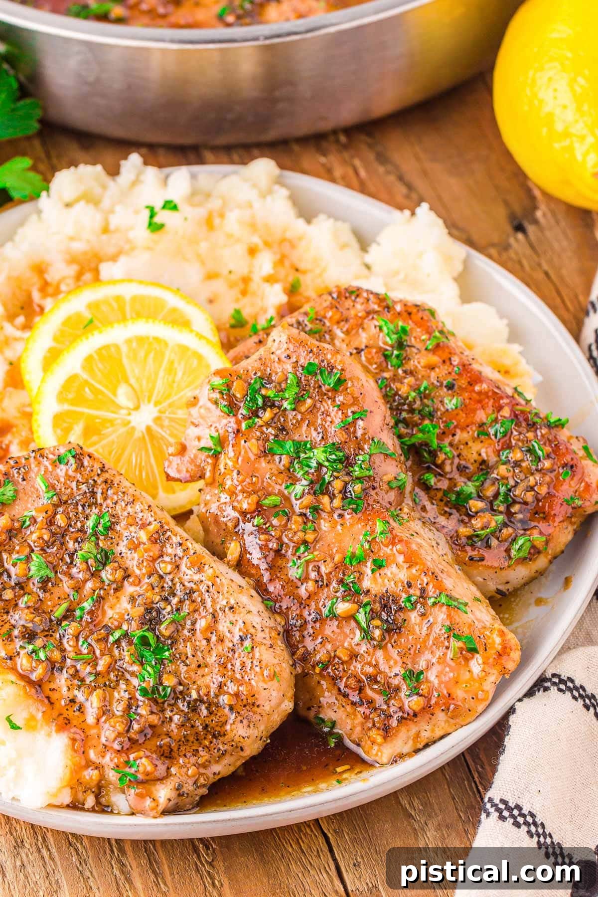 Three golden-brown honey garlic pork chops resting on a bed of creamy mashed potatoes, garnished with fresh parsley and vibrant lemon slices. The plate is presented on a rustic wooden board, emphasizing a comforting and appealing meal.