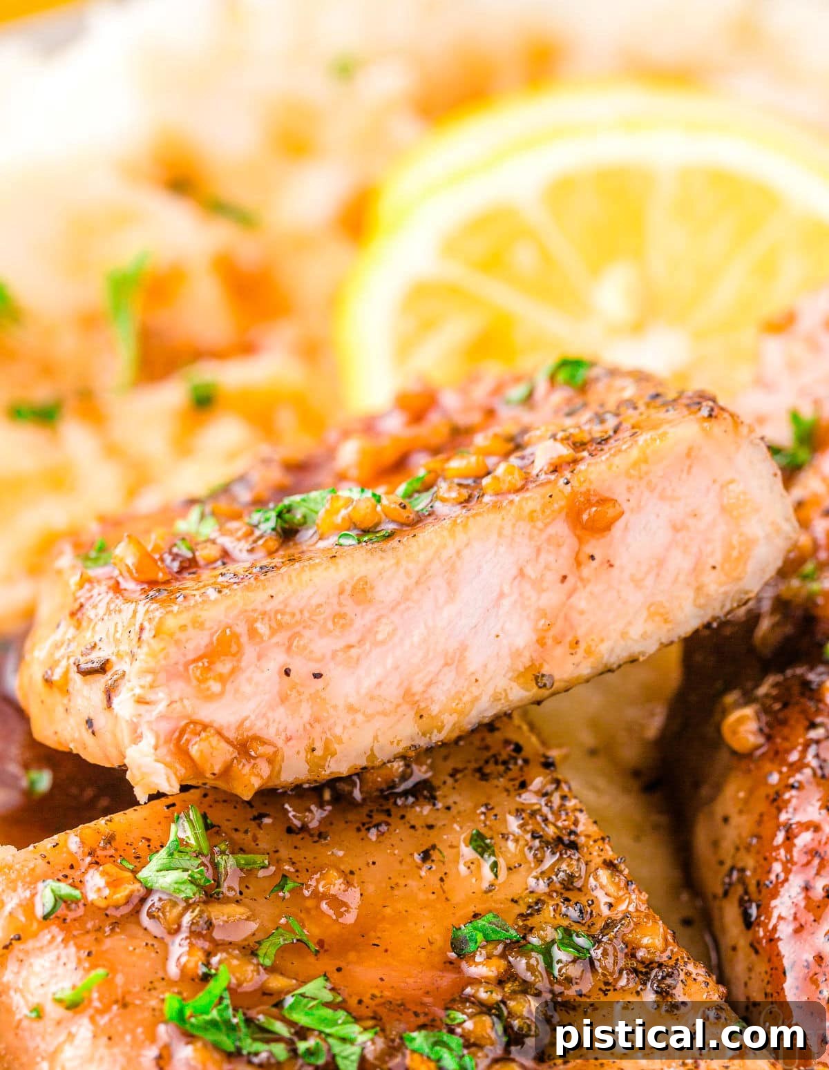 A perfectly cooked honey garlic pork chop, sliced in half to reveal its juicy interior, with the luscious glaze generously dripping down its sides. Fresh parsley garnish and blurred lemon slices add a touch of freshness and elegance to the background.