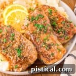 Three honey garlic pork chops sitting on a bed of mashed potatoes topped with fresh parsley and lemon slices. plate is on a wood board.