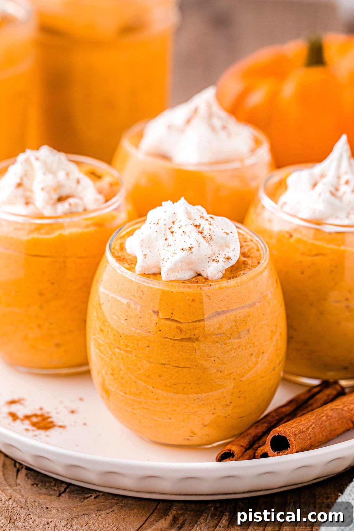Four elegant dessert glasses filled with creamy pumpkin mousse, topped with a swirl of whipped cream and a sprinkle of pumpkin pie spice. Two cinnamon sticks rest on the white plate holding the glasses, all on a warm wood surface, creating a perfect autumn scene.