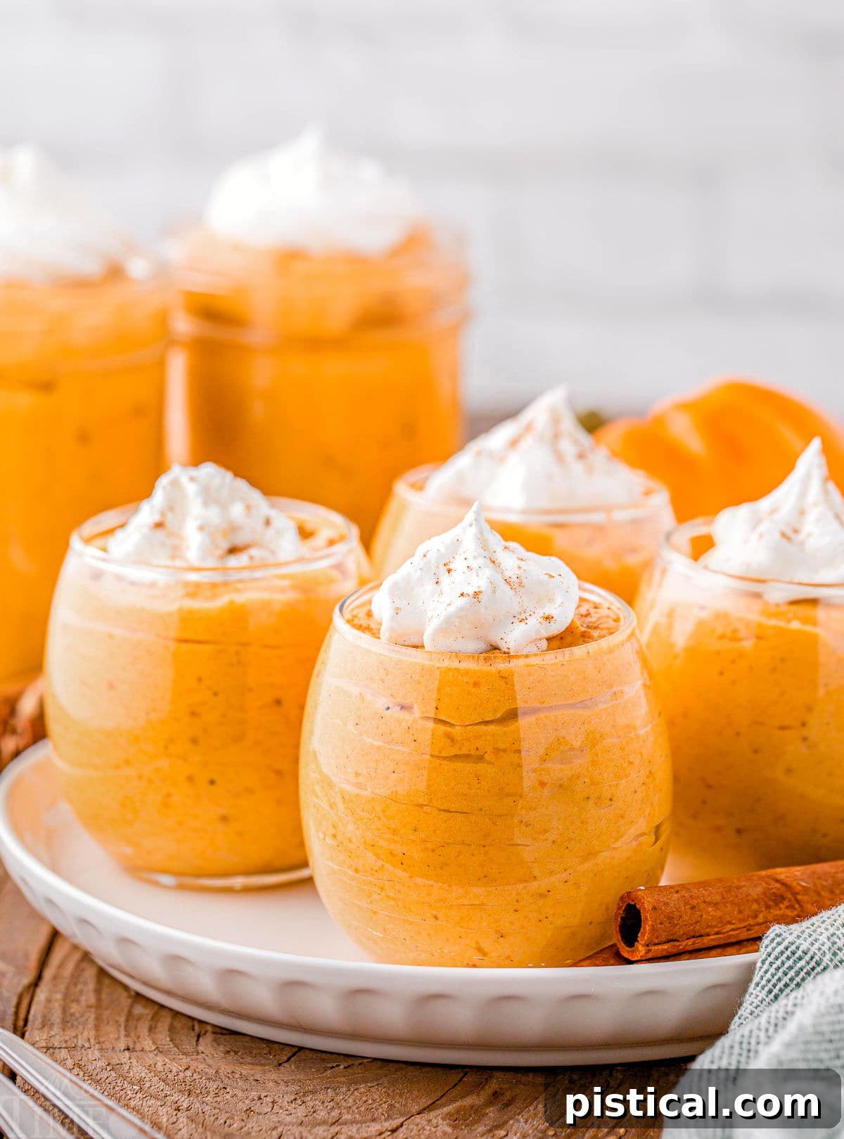 Four short wine glasses filled with pumpkin mousse, topped with whipped cream and pumpkin pie spice. The glasses are arranged on a round white plate on a wood surface, with two cinnamon sticks beside them for a rustic touch.