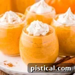 Four stemless wine glasses filled with pumpkin mousse, topped with whipped cream and pumpkin pie spice. The glasses are arranged on a round white plate on a wood surface, with two cinnamon sticks for decoration.