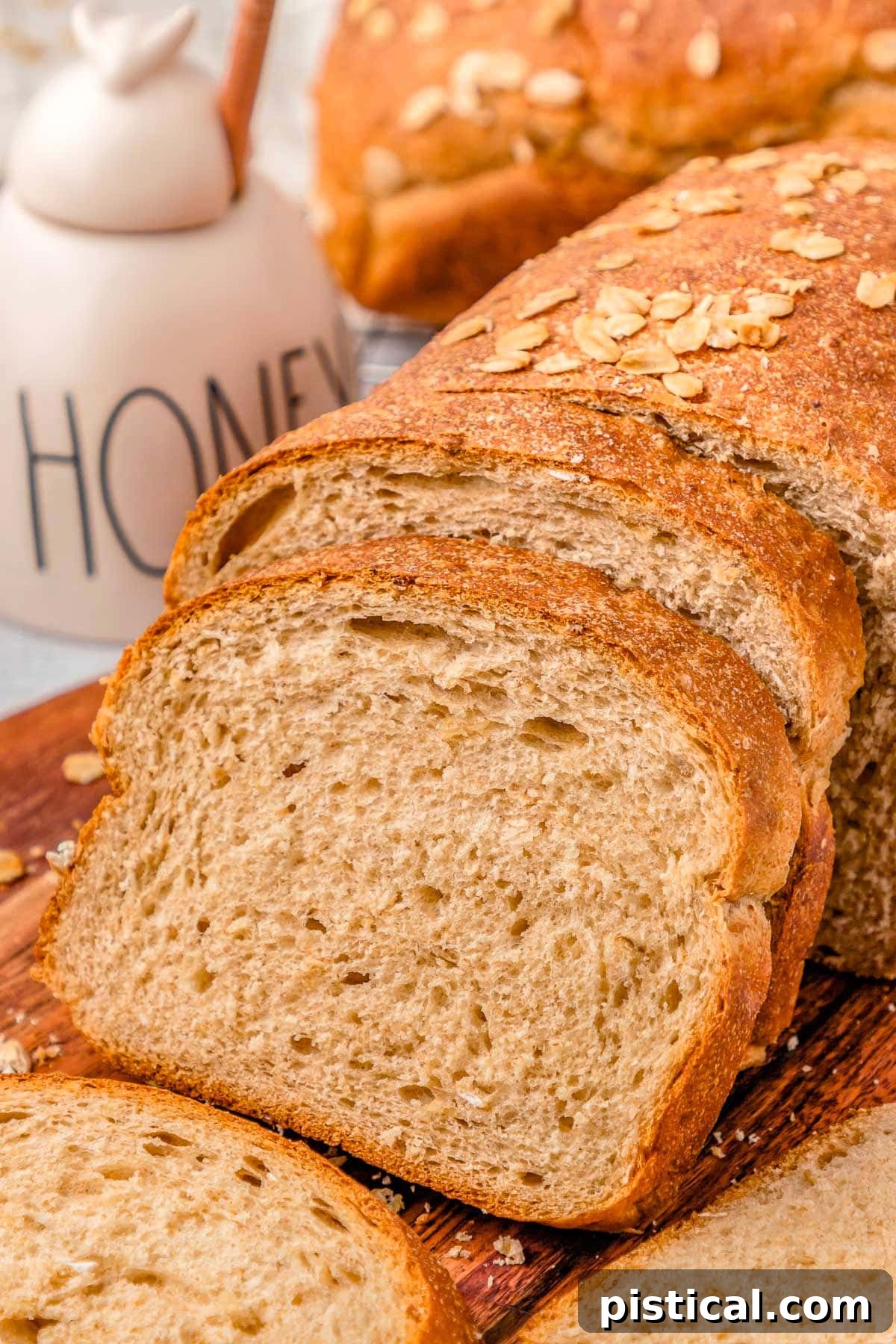 two slices cut off a loaf of wheat bread on a wood board. more bread can be seen in the background next to a white honey jar.