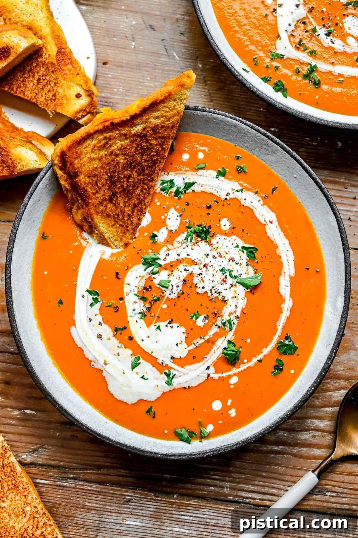 A top-down view of a cozy bowl of Instant Pot Tomato Soup, featuring half of a classic grilled cheese sandwich dipped into the rich, creamy liquid. Garnished with a swirl of heavy cream, fresh basil, and a sprinkle of black pepper.