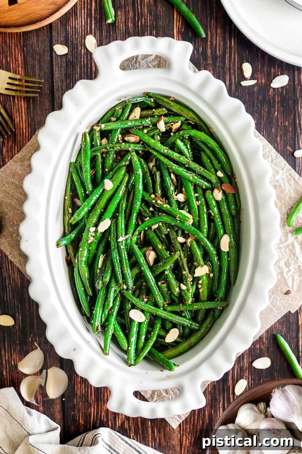 Large white oval serving bowl filled with bright green garlic green beans, garnished with toasted almonds, on a dark wood surface. A top-down view highlighting the vibrant colors and appealing texture.
