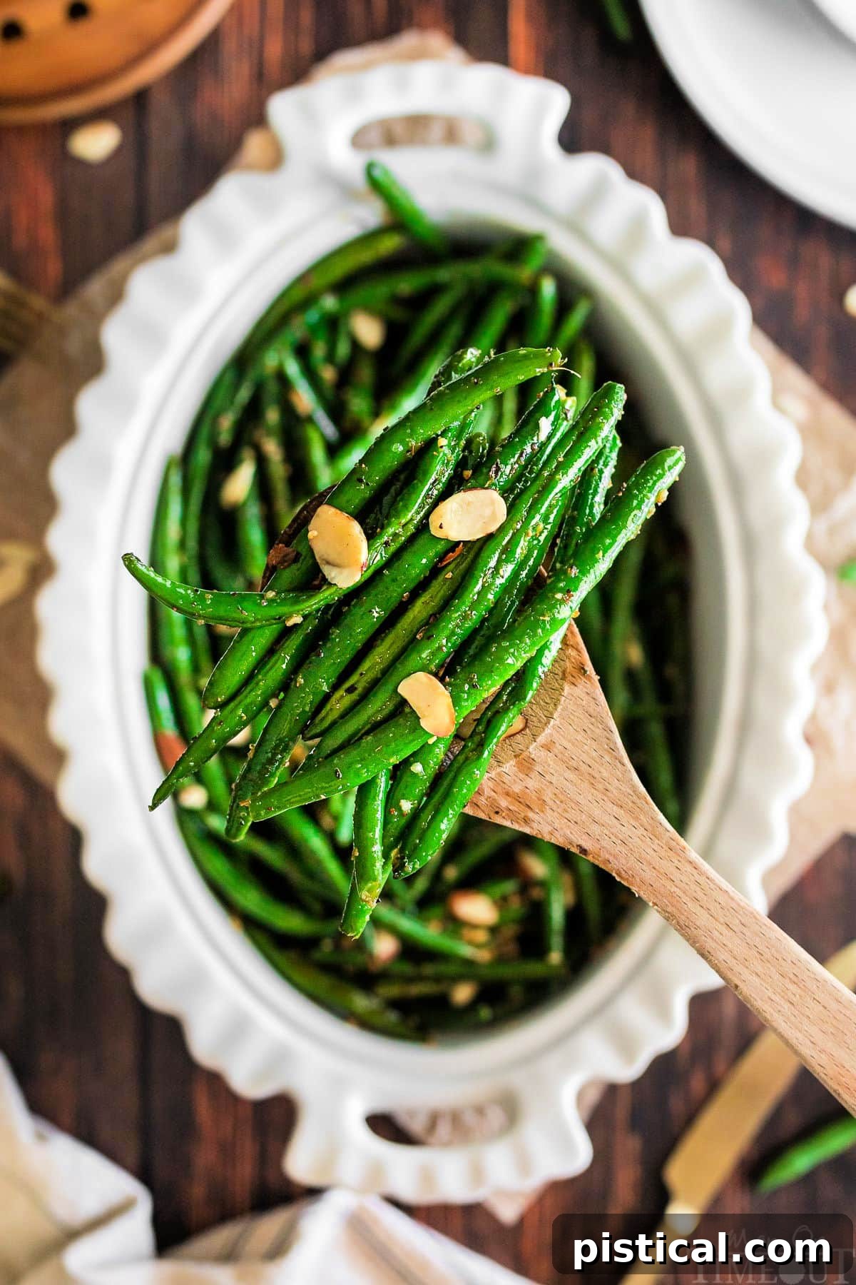 A wooden spoon lifting a serving of brown butter garlic green beans from an oval white serving dish, highlighting the inviting presentation.