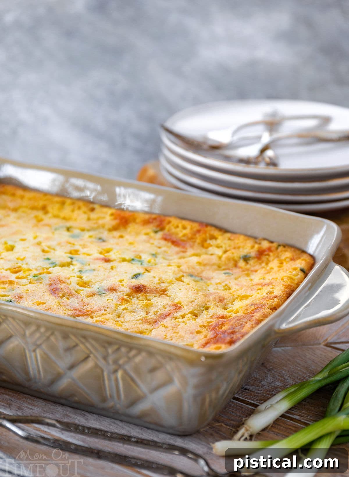 Close-up of a delicious corn casserole in a brown baking dish, garnished with fresh green onions, ready for serving. Stacked plates are visible in the background.