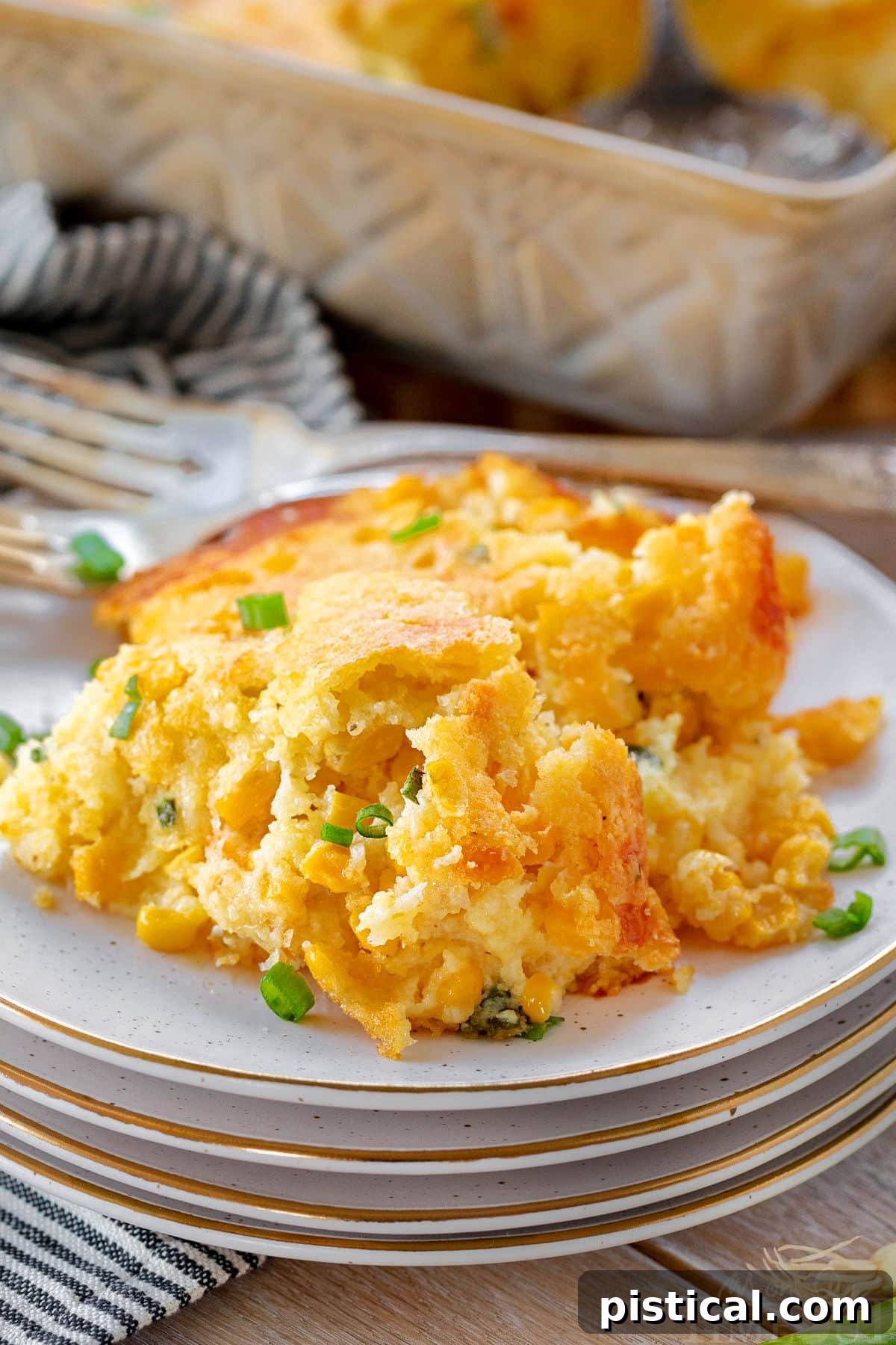 A scoop of rich and creamy corn casserole on a stack of plates, with fresh green onions sprinkled on top. The main casserole dish is visible in the background.
