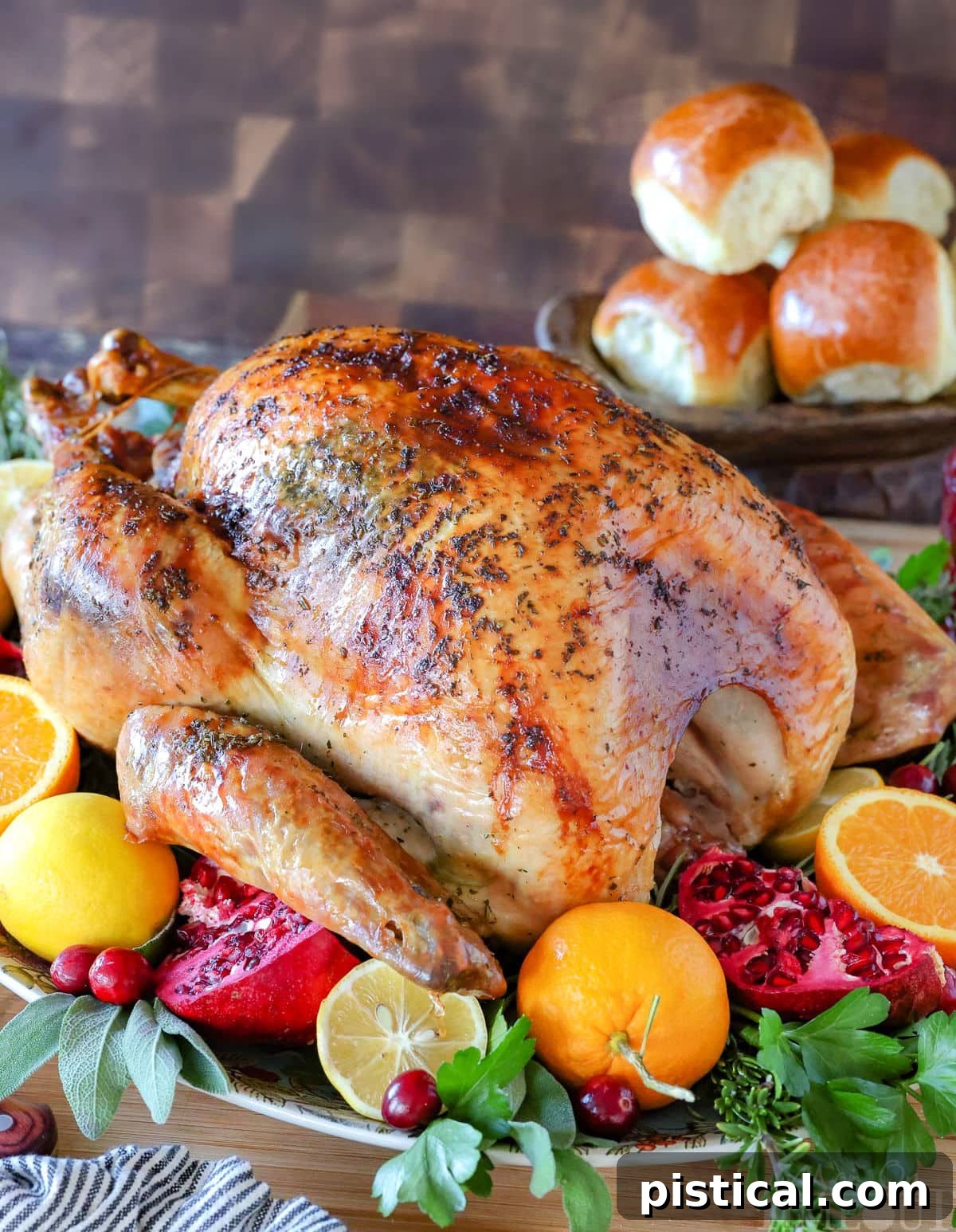 A perfectly roasted whole Thanksgiving turkey, garnished with fresh fruit and herbs, presented on a serving platter. Dinner rolls are visible in the background.