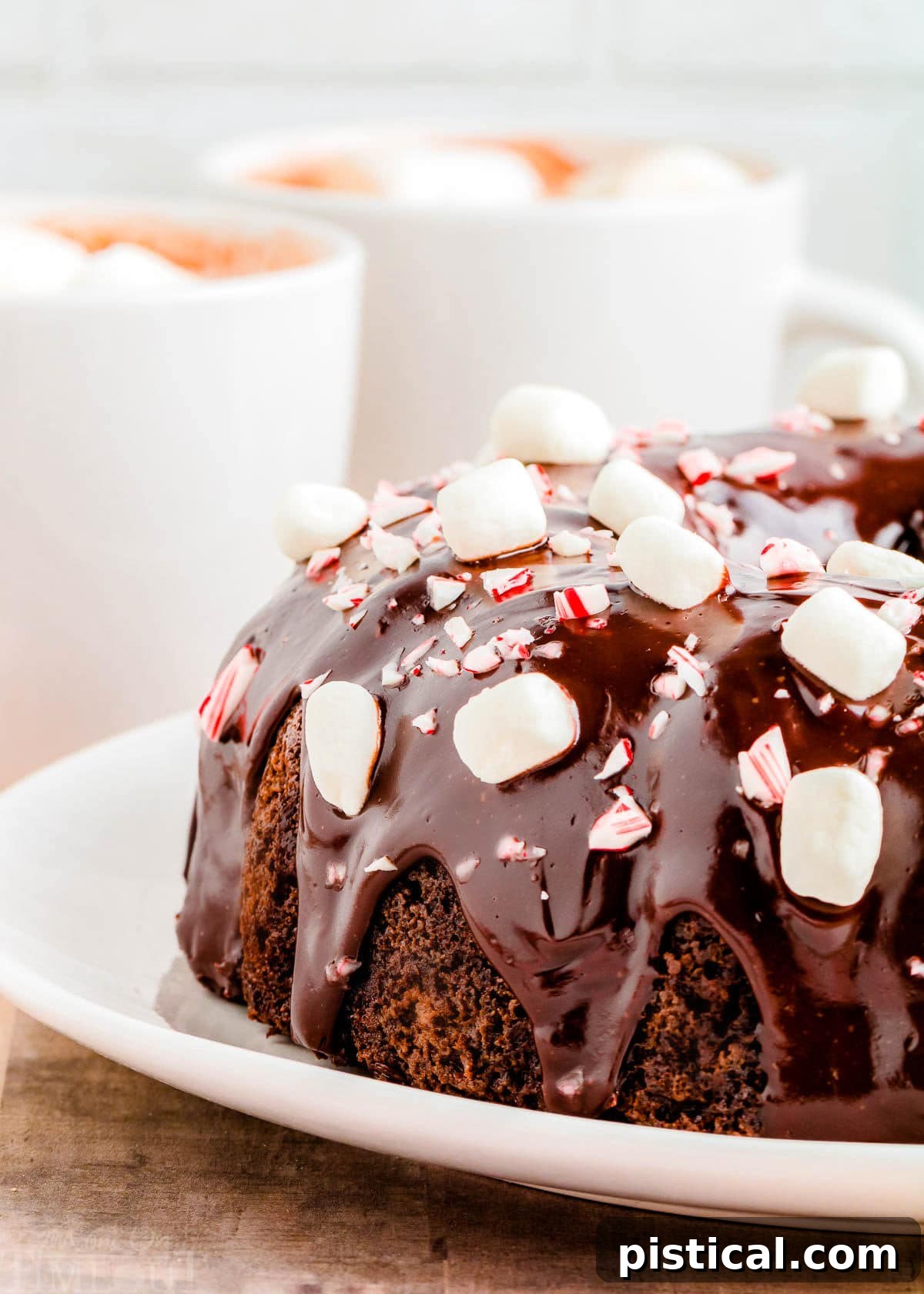 peppermint hot cocoa cake on a white plat decorated with miniature marshmallows and crushed candy canes. two mugs of hot cocoa can be seen in the background.