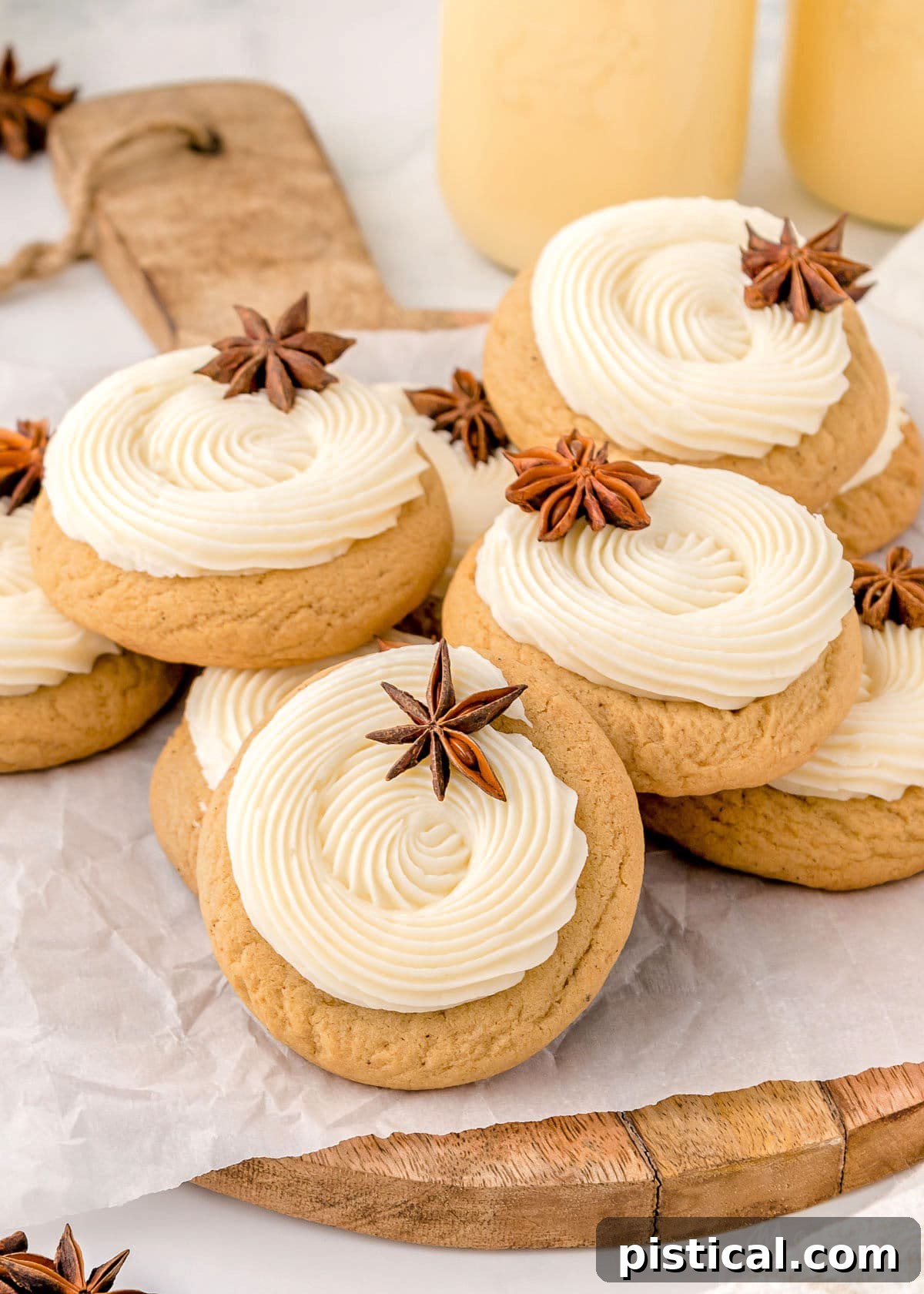 A charming stack of freshly baked eggnog cookies on a rustic wooden cutting board. Each cookie is generously topped with a thick swirl of creamy eggnog frosting and delicately garnished with a star anise, creating a beautiful and aromatic presentation. Two inviting cups of eggnog sit blurred in the background, hinting at the perfect holiday pairing.