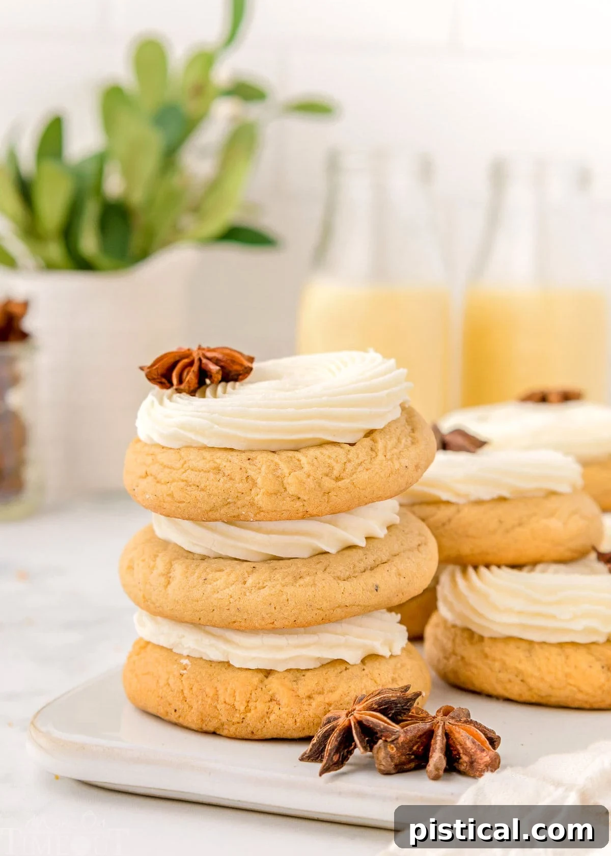 Three perfectly frosted eggnog cookies are elegantly stacked on a clean white board, showcasing their inviting texture and rich frosting. More of these festive cookies are artfully arranged in the background, creating an enticing display of holiday baking perfection.