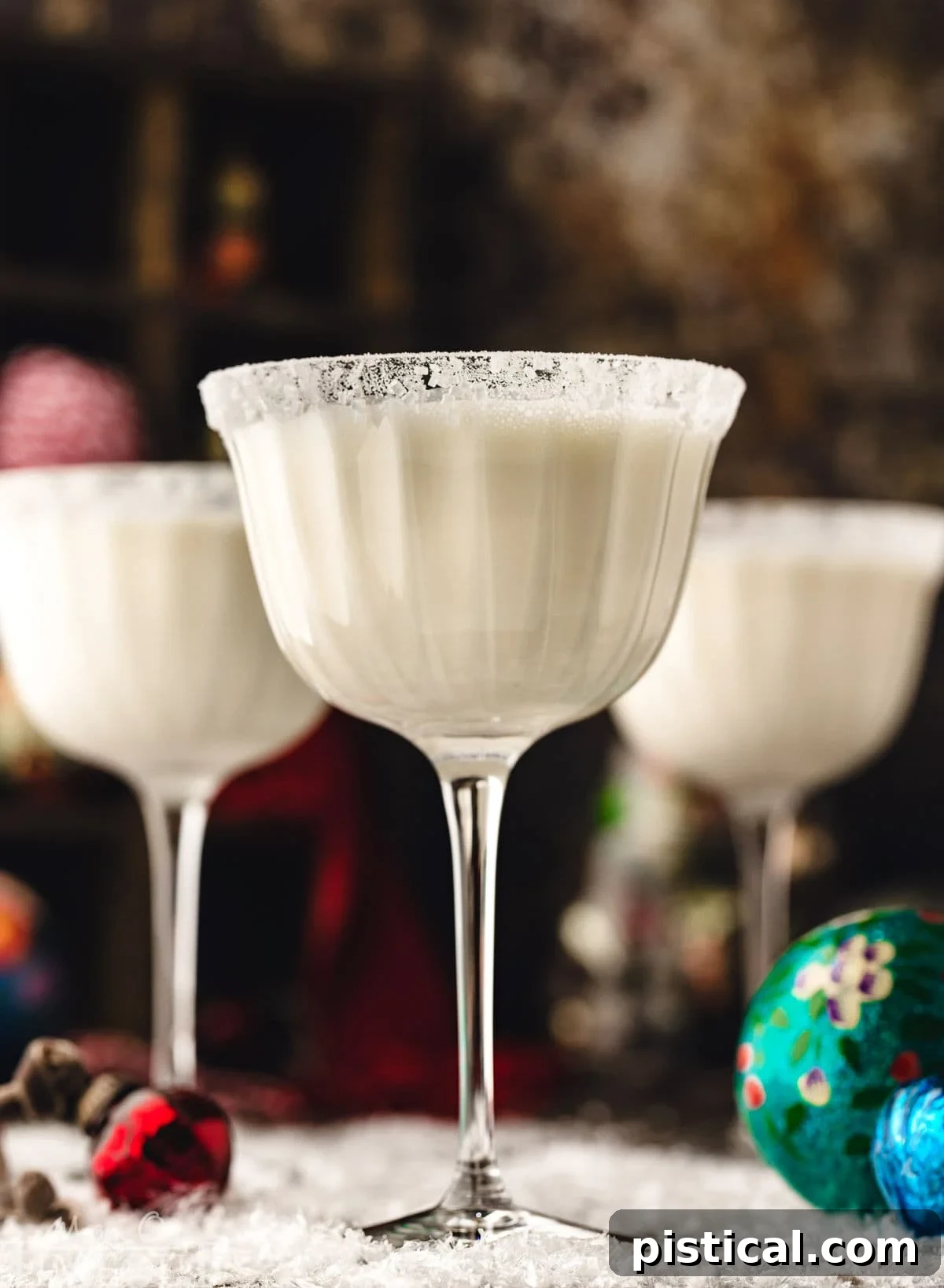 three white chocolate martinis in tall glasses dusted with powdered sugar for a snowy look. the cocktails are sitting on a snowy surface with glass ornaments scattered about.