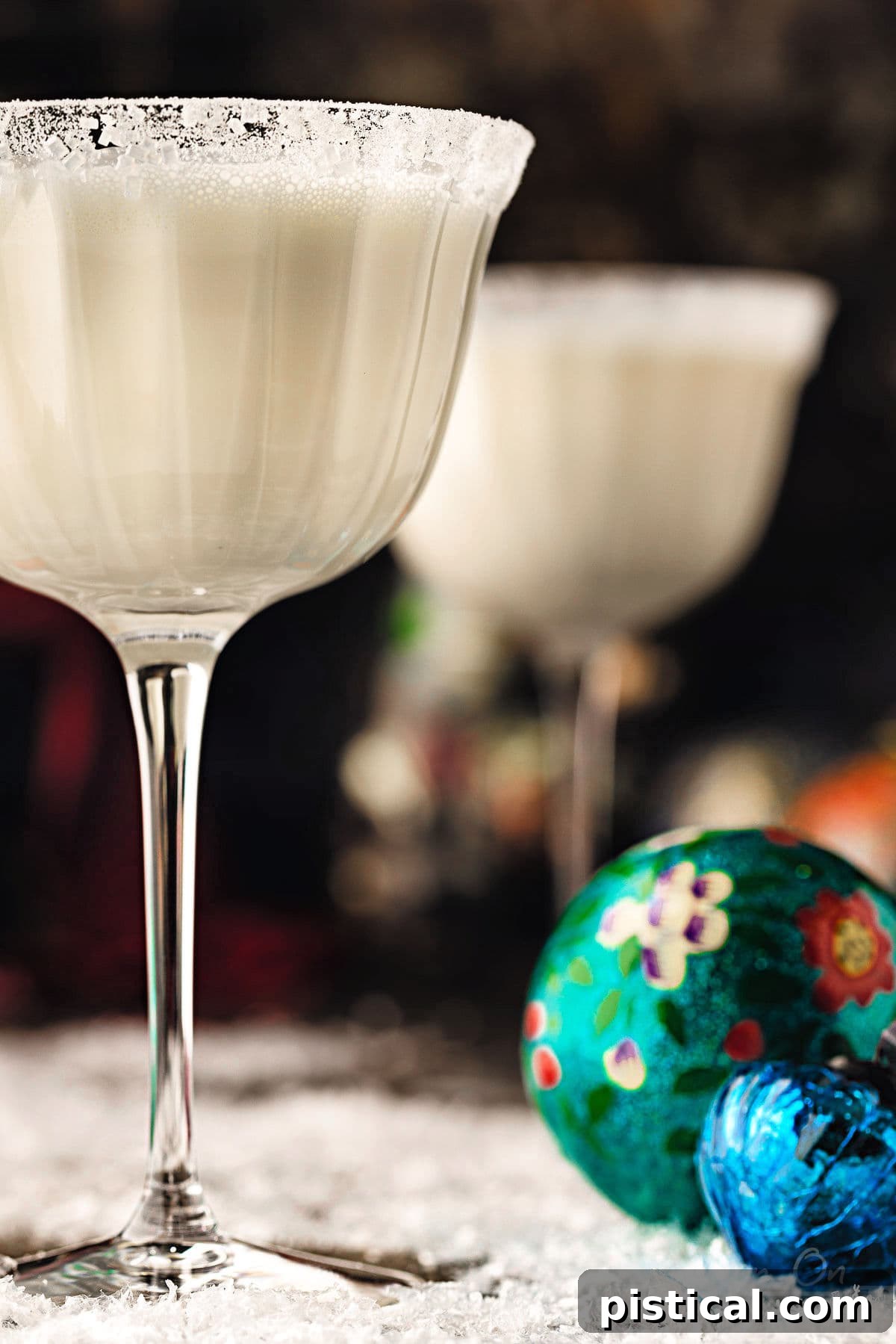 two white martinis in tall glass dusted with powdered sugar for a snowy look. the cocktails is sitting next to vintage ornaments. bottles of vodka and white chocolate liqueur can be seen in the background.