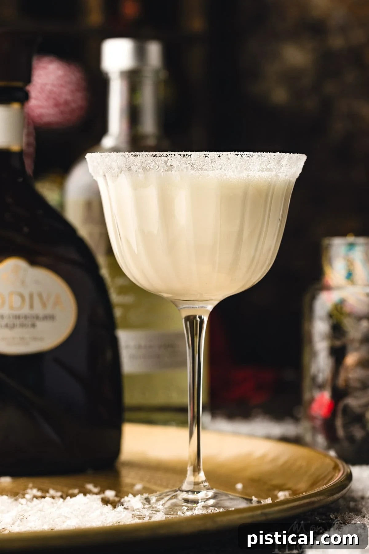 white chocolate martini cocktail in tall glass dusted with powdered sugar for a snowy look. the cocktails is sitting on a gold tray. bottles of vodka and white chocolate liqueur can be seen in the background.