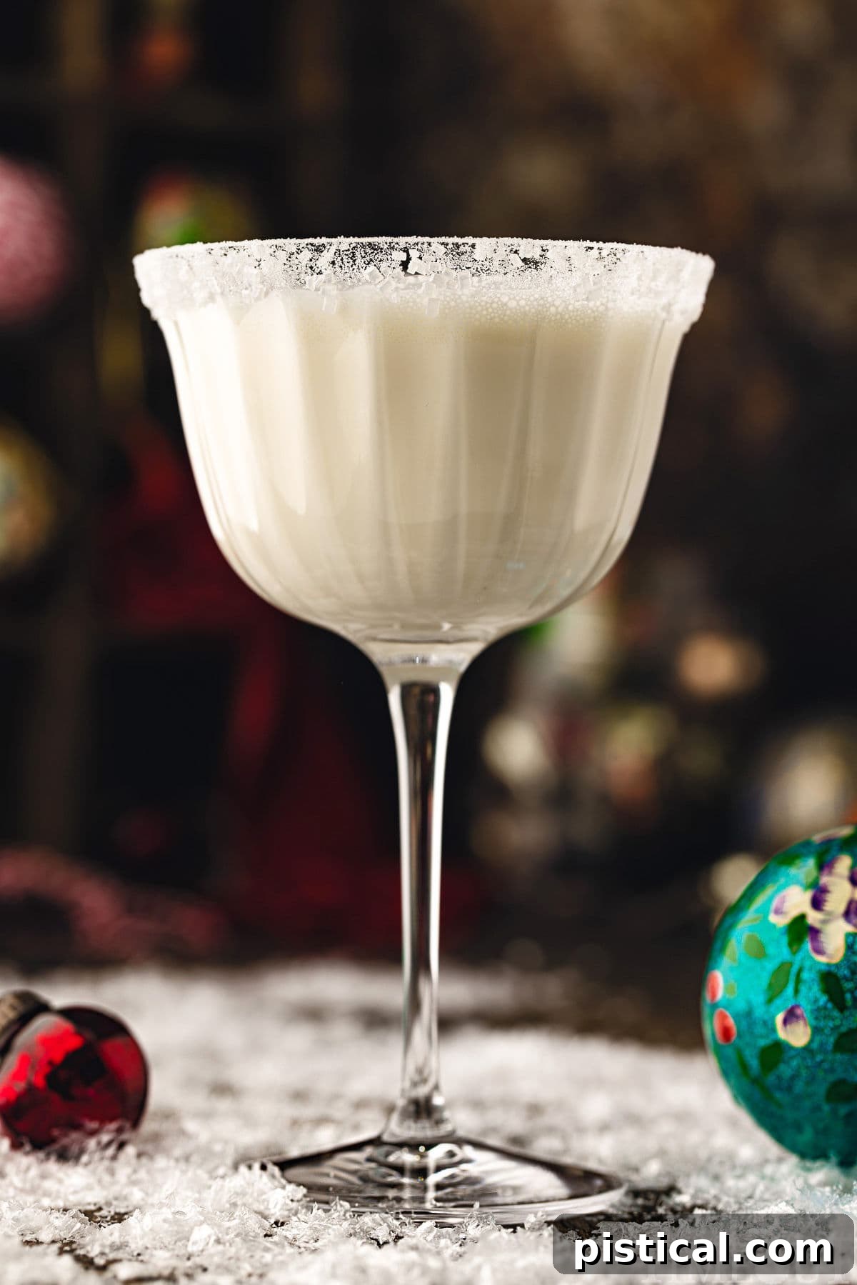 white chocolate martini cocktail in tall glass dusted with powdered sugar for a snowy look. the cocktails is sitting on a snowy surface with glass ornaments scattered about.