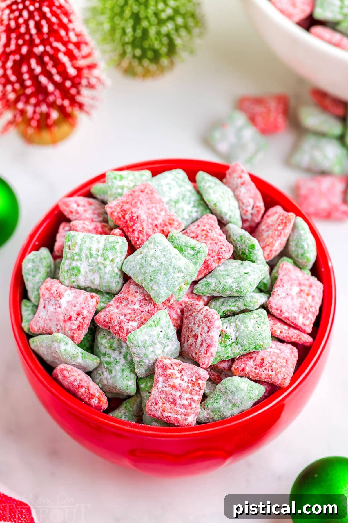 red bowl filled with red and green puppy chow. the bowl is on a white surface which christmas decorations in the background.