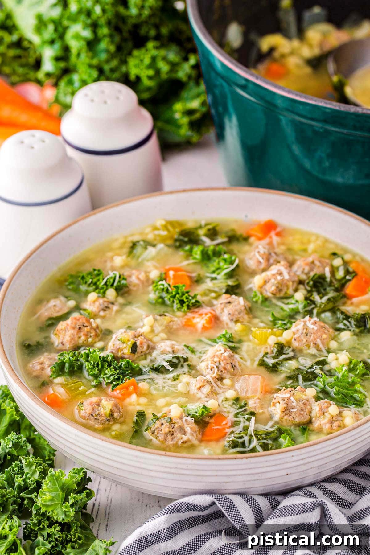 large bowl filled with Italian Wedding Soup and garnished with Parmesan cheese. A dark teal dutch oven can be seen in the background. A black and white striped towel is in the front of the bowl.