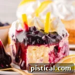 slice of lemon blueberry cheesecake topped with blueberry pie filling, whipped cream and two lemon slices sitting on small round plate with one bite taken from the top if the slice.