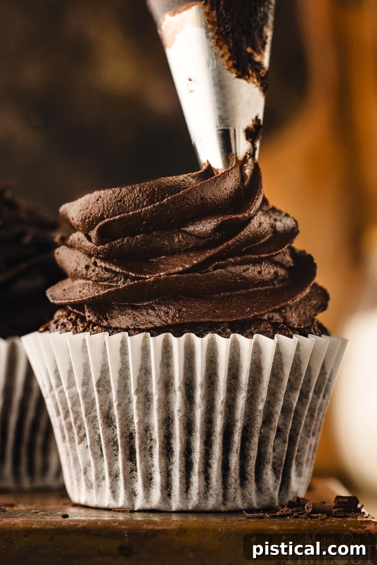 Chocolate buttercream being expertly piped onto a freshly baked chocolate cupcake, which rests on a dark metal surface. The frosting forms elegant swirls, showcasing its smooth and rich texture.