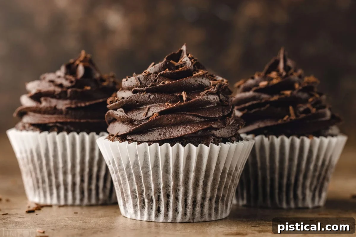 Three perfectly baked chocolate cupcakes arranged in a neat row, each nestled in a white parchment paper liner and generously topped with expertly piped chocolate buttercream. The inviting visual showcases their readiness for enjoyment.