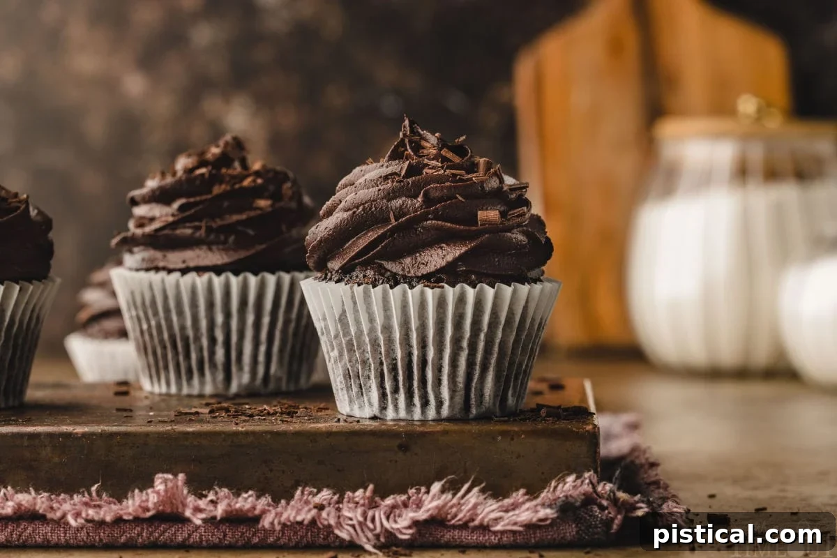 Two perfectly baked chocolate cupcakes, presented in crisp white liners and topped with abundant swirls of chocolate buttercream. A cutting board in the soft background hints at a homemade kitchen setting.