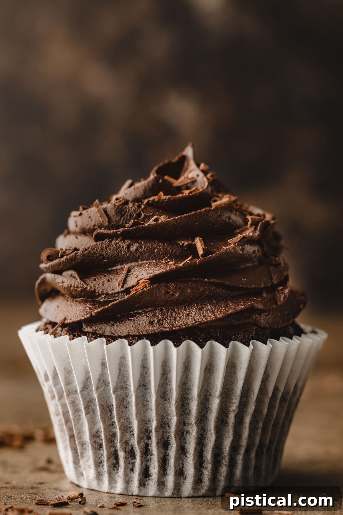 A perfect chocolate cupcake, still in its crisp white wrapper, is topped with a generous swirl of deep chocolate buttercream. The background is dark and moody, emphasizing the rich color and inviting texture of the cupcake.