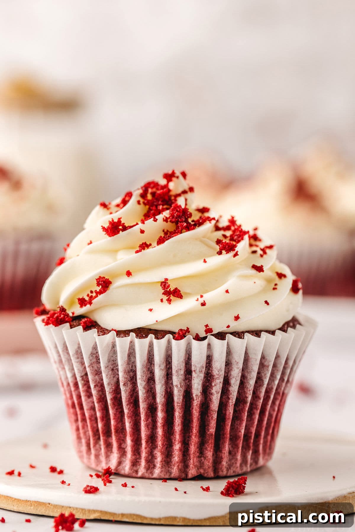 A close-up shot of a perfect Red Velvet Cupcake nestled in a white liner on a white pottery plate. Its cream cheese frosting is piped into an elegant swirl, garnished with red velvet crumbs. Several other frosted cupcakes blur in the background, hinting at a delightful spread.