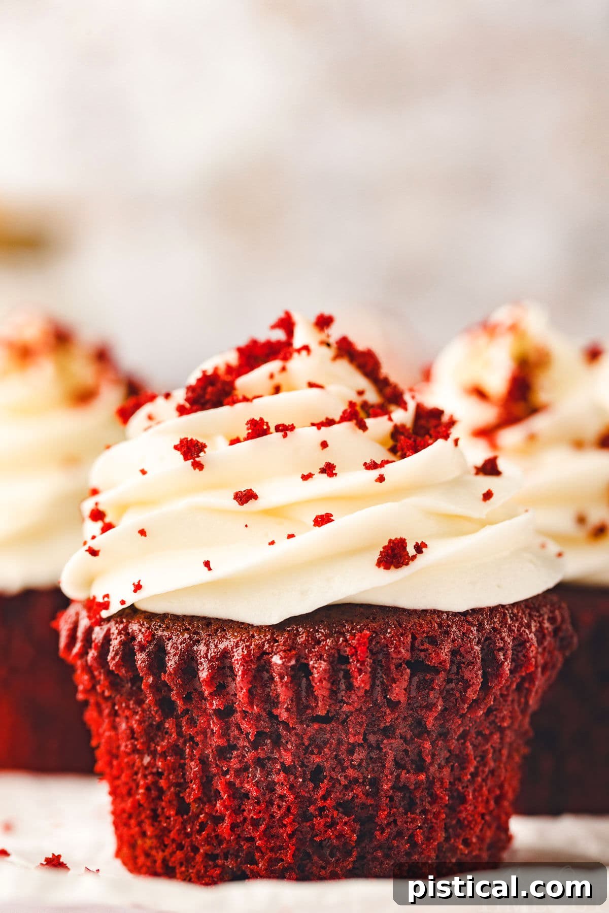 A beautifully composed shot featuring a single Red Velvet Cupcake on a white pottery plate, crowned with a generous swirl of cream cheese frosting and a scattering of red velvet crumbs. In the soft-focus background, more cupcakes promise further delight.