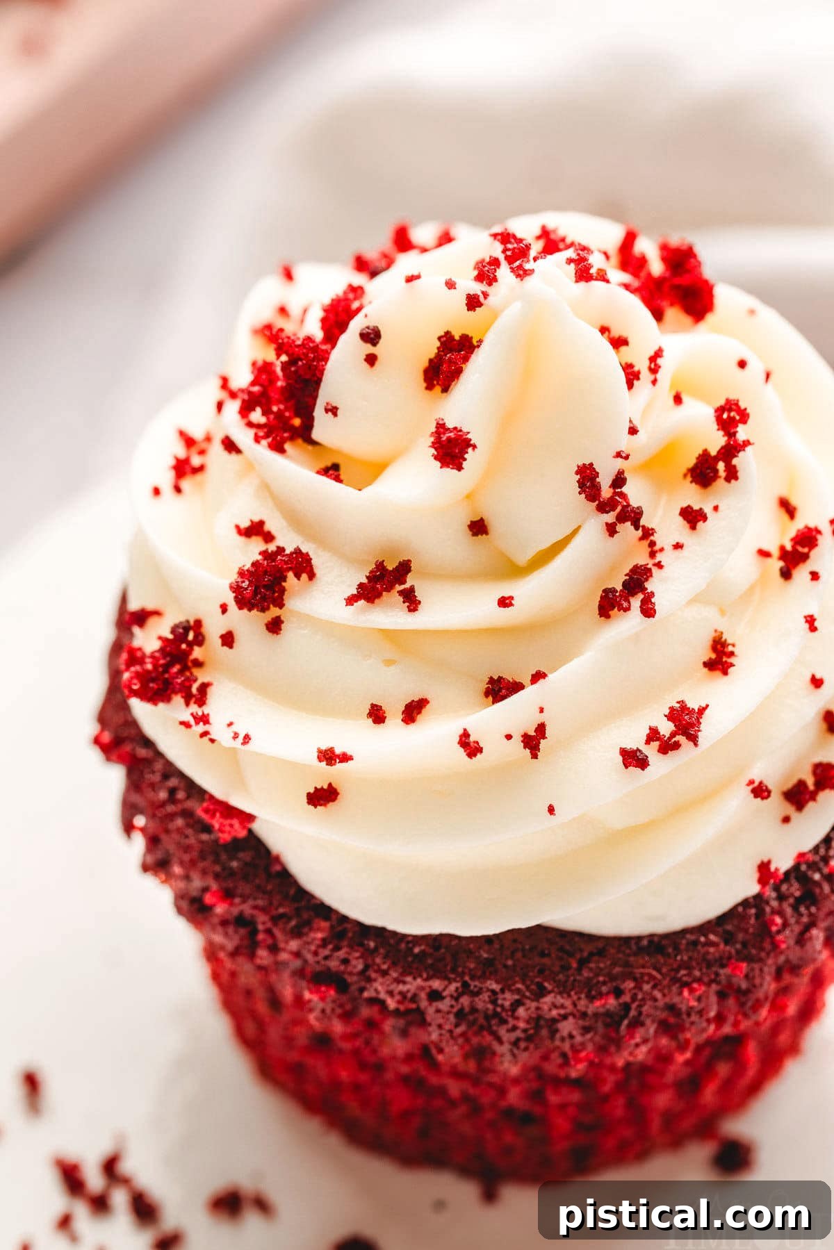 A top-down, close-up view of a Red Velvet Cupcake, featuring a luxurious swirl of creamy white cream cheese frosting. The frosting is beautifully adorned with fine red velvet crumbs, creating an appealing texture and color contrast.