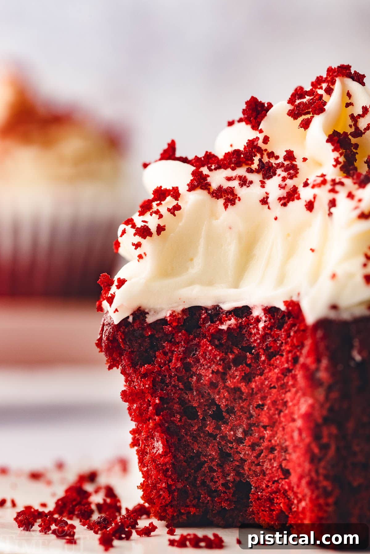 A single red velvet cupcake, exquisitely frosted with cream cheese frosting, is shown in close-up. A bite has been taken, revealing the wonderfully vibrant red interior and its moist texture.