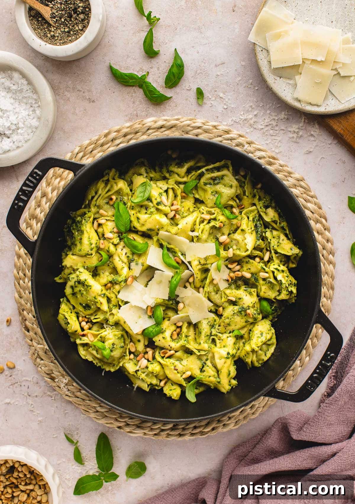 Top-down shot of a Pesto Tortellini recipe in a large black cast iron skillet, elegantly garnished with shaved Parmesan, toasted pine nuts, and fresh basil leaves. Surrounding the skillet are small bowls containing Parmesan, salt, and pepper.