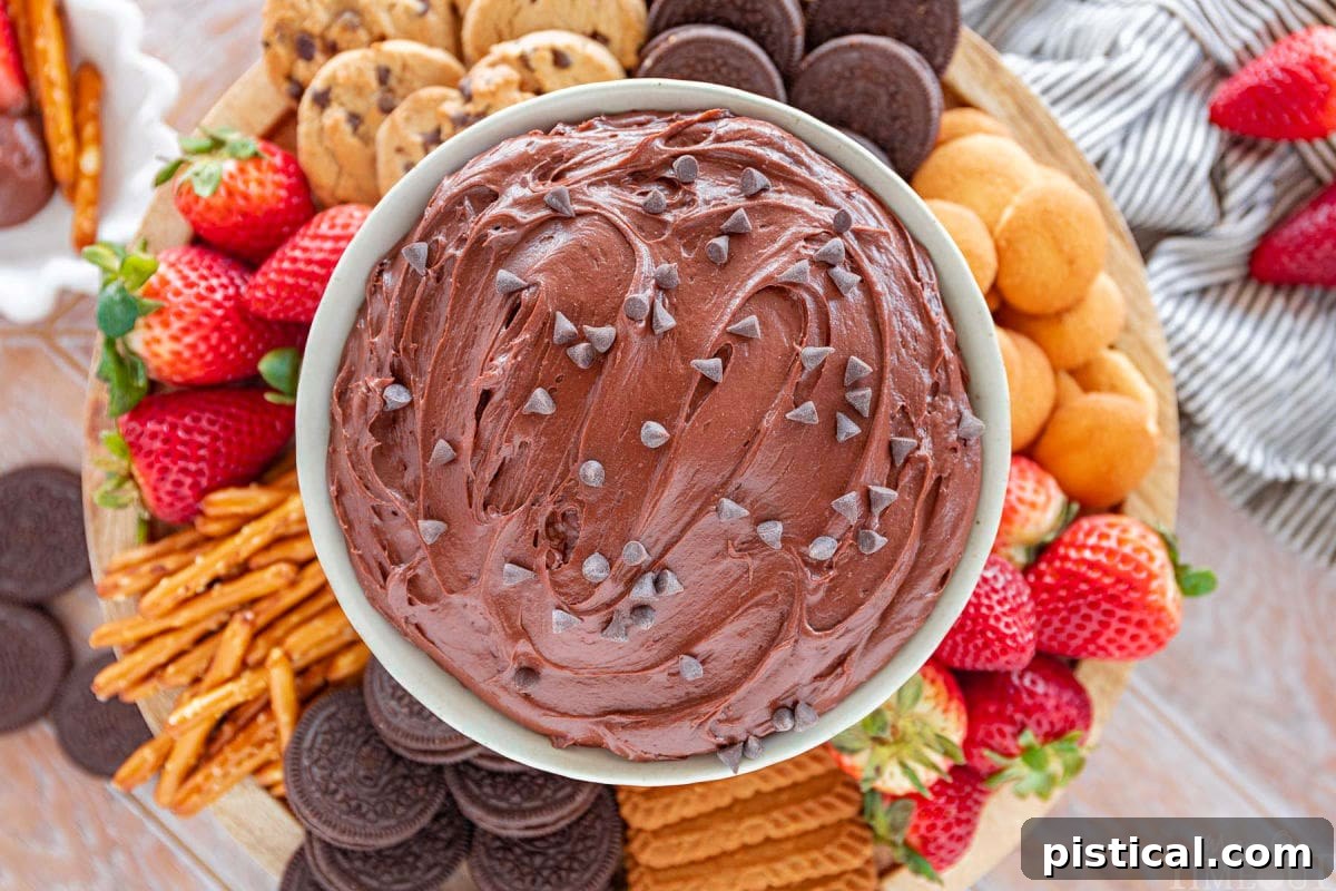 wide shot of top down look at edible brownie batter in serving bowl topped with mini chocolate chips surrounded by strawberries, pretzels, cookies and more dippers.