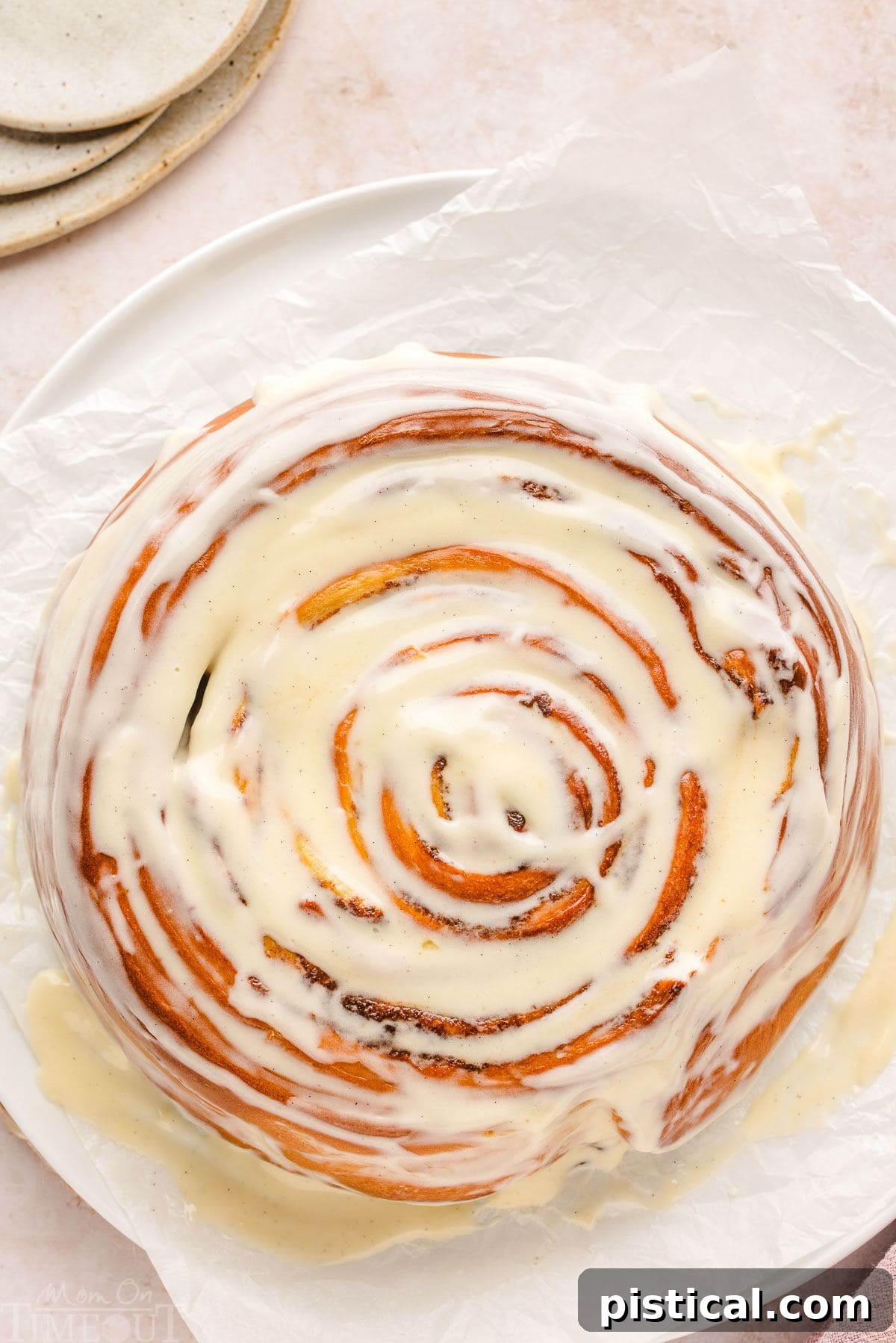A large, perfectly round Giant Cinnamon Roll Cake, generously covered in a luscious cream cheese glaze, resting on white parchment paper. Small white plates are set to the side, ready for serving.