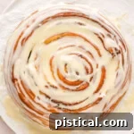 Top-down view of a giant cinnamon roll cake sitting on a white piece of parchment paper with a cream cheese glaze generously drizzled over the top.