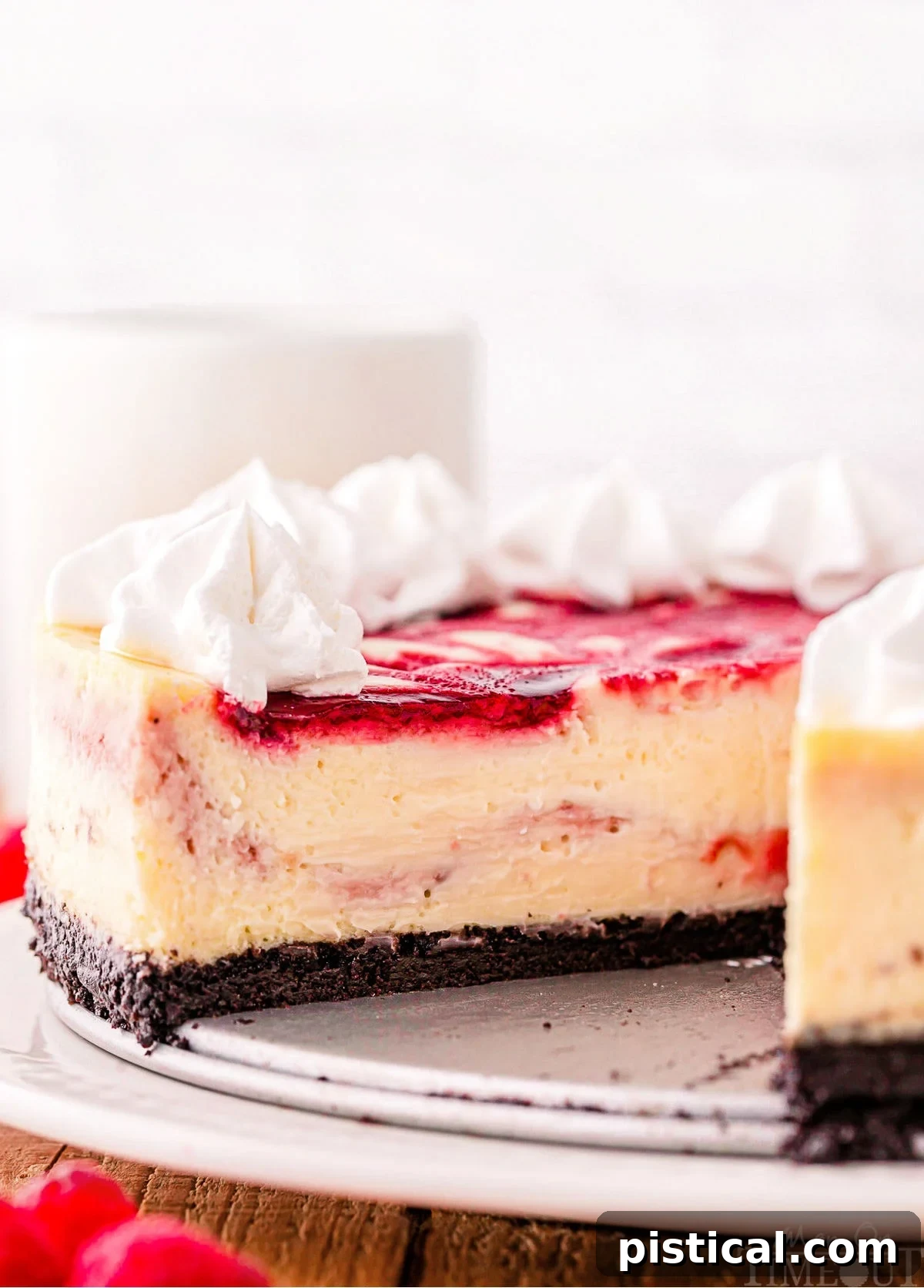 A profile view of a tall white chocolate raspberry cheesecake, showcasing its thick OREO crust and a generous topping of whipped cream.