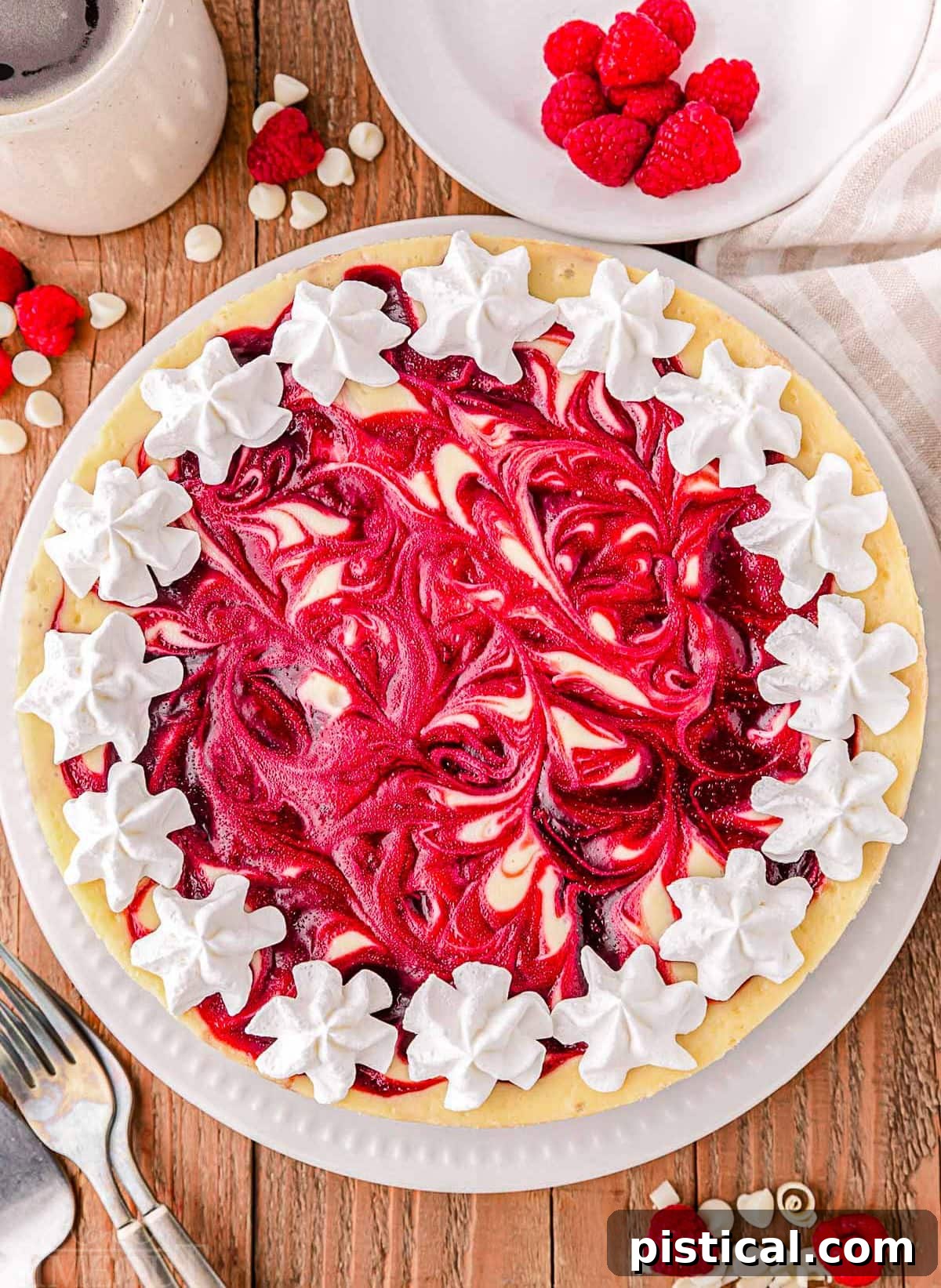 A top-down view of a whole white chocolate raspberry cheesecake, elegantly topped with a raspberry swirl and garnished with delicate dollops of fresh whipped cream and whole raspberries.
